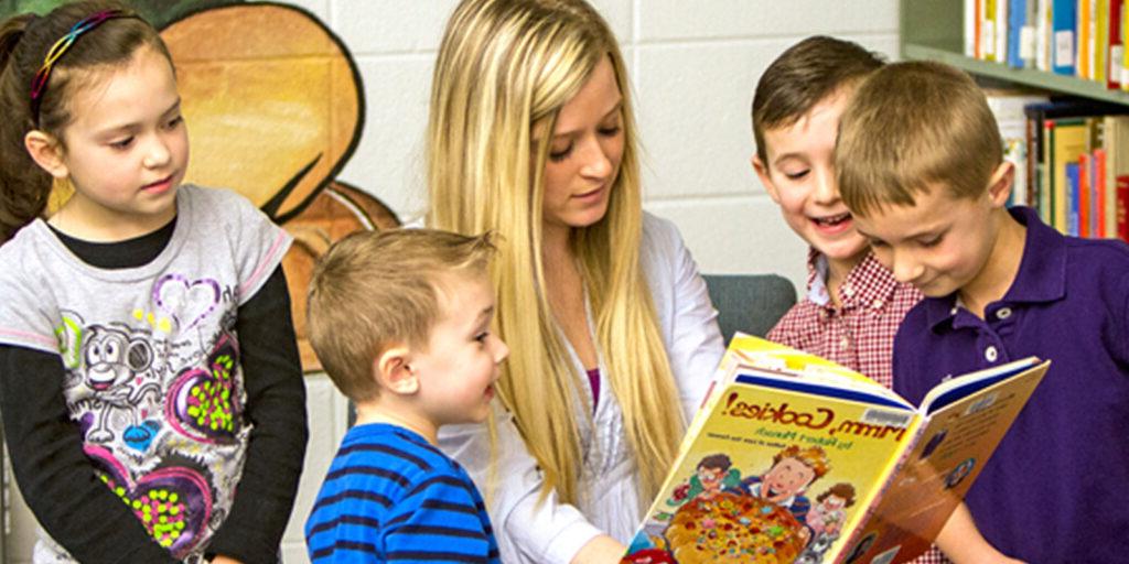 A young woman is reading a book titled "Warm Cookies!" to four young children, who are gathered closely around her, in a library setting.