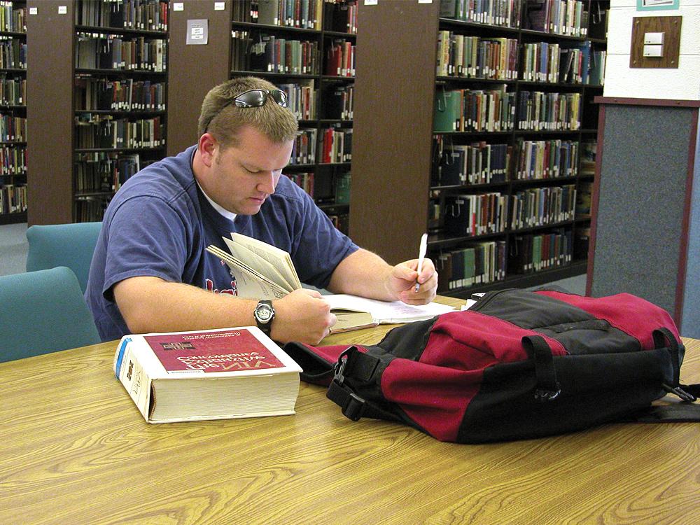 A person wearing a blue shirt is studying at a library table with a red and black backpack and an open book in front of them. Bookshelves are visible in the background.